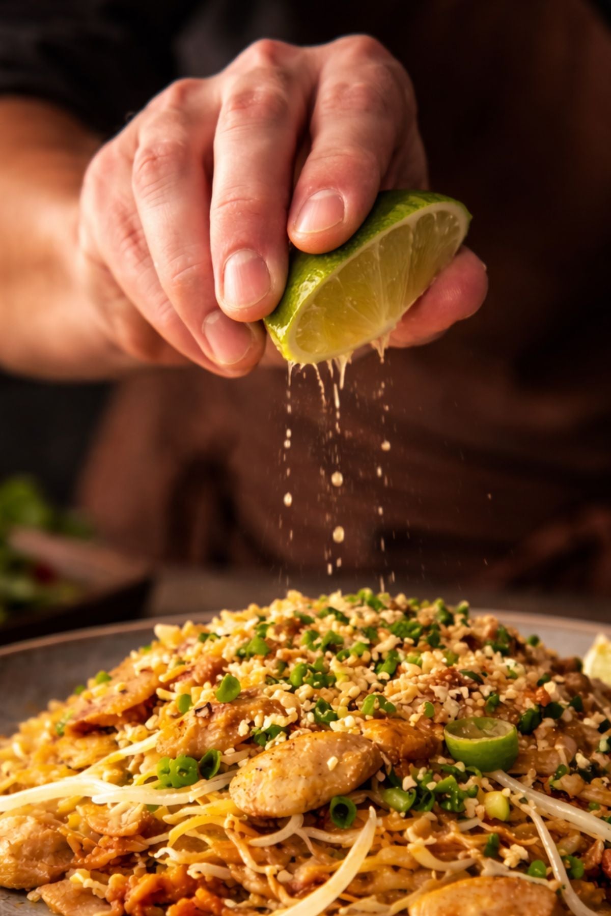 Eine Hand drückt Limette über Nudeln mit Erdnüssen, Frühlingszwiebeln und Hühnchen, angerichtet in einer Fissler-Edelstahl-Kochschüssel.