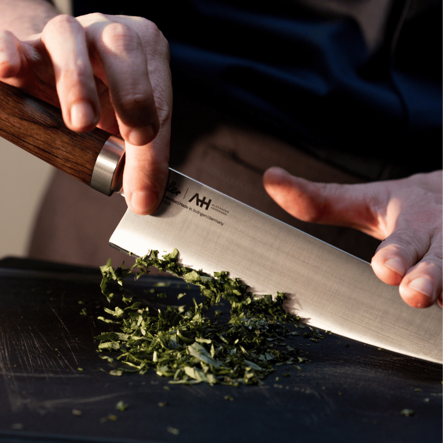 Eine Person hackt fachmännisch frische Kräuter mit einem Präzisionskochmesser von Fissler auf einem schwarzen Schneidebrett, das deutsche Handwerkskunst demonstriert.