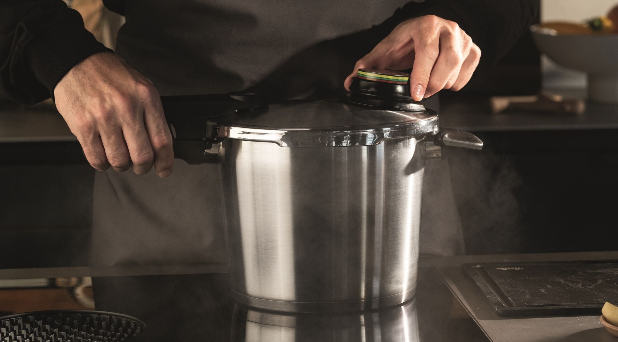 Eine Person öffnet einen dampfenden Fissler-Edelstahl-Schnellkochtopf auf einer Herdplatte, der deutsche Präzision und zeitloses Design unterstreicht.
