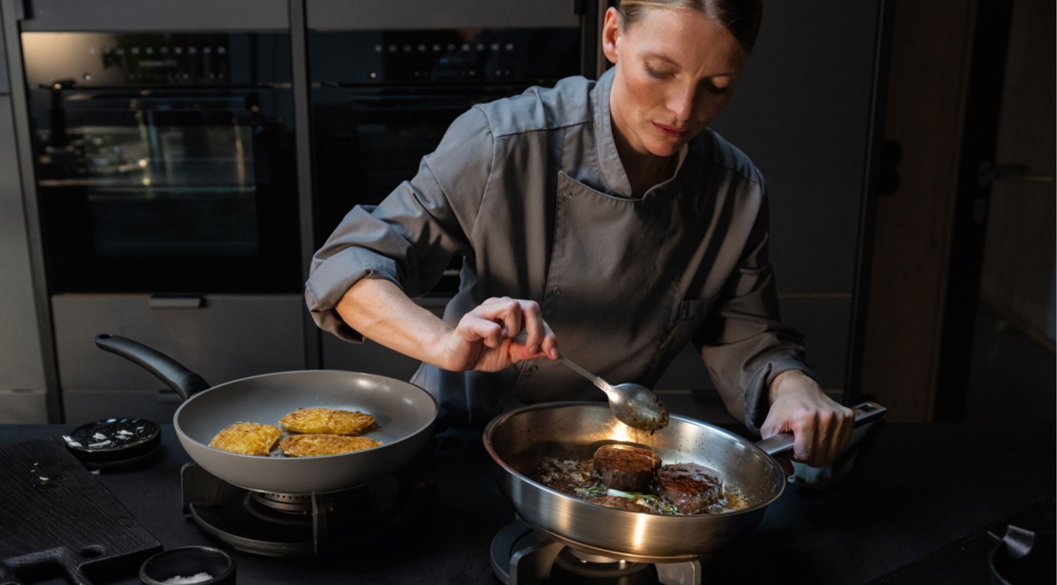 Ein Koch in grauer Uniform kocht mit Fissler-Edelstahlpfannen und löffelt Soße über Fleisch auf dem Herd in einer eleganten modernen Küche.