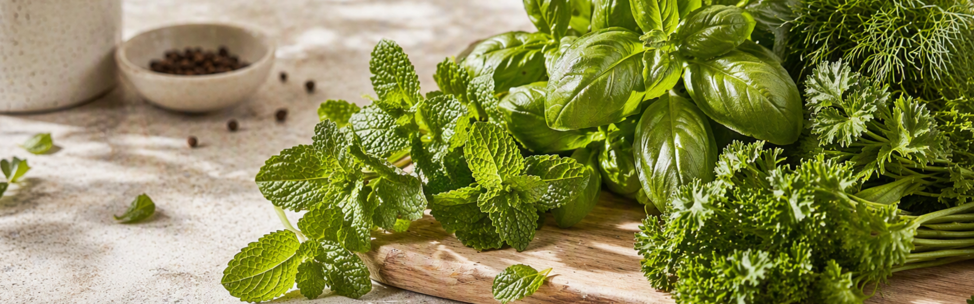 Fresh green herbs—mint, basil, dill, and parsley—are artfully arranged on a wooden cutting board set alongside peppercorns and a cup. The scene evokes Fissler’s commitment to timeless design, precision engineering, and high-quality German craftsmanship found in our renowned cookware collections.
