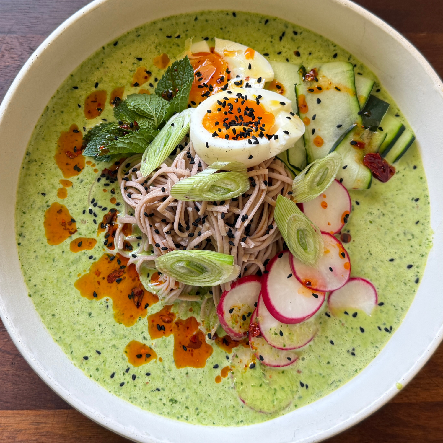 A beautifully arranged bowl of green broth with soba noodles, sliced radish, cucumber, scallions, fresh mint, a halved soft-boiled egg, black sesame seeds and chili oil. The visually striking dish is presented in a way that highlights attention to detail—crafted with tools like Fissler’s precision-engineered cookware for optimal cooking results and timeless design.