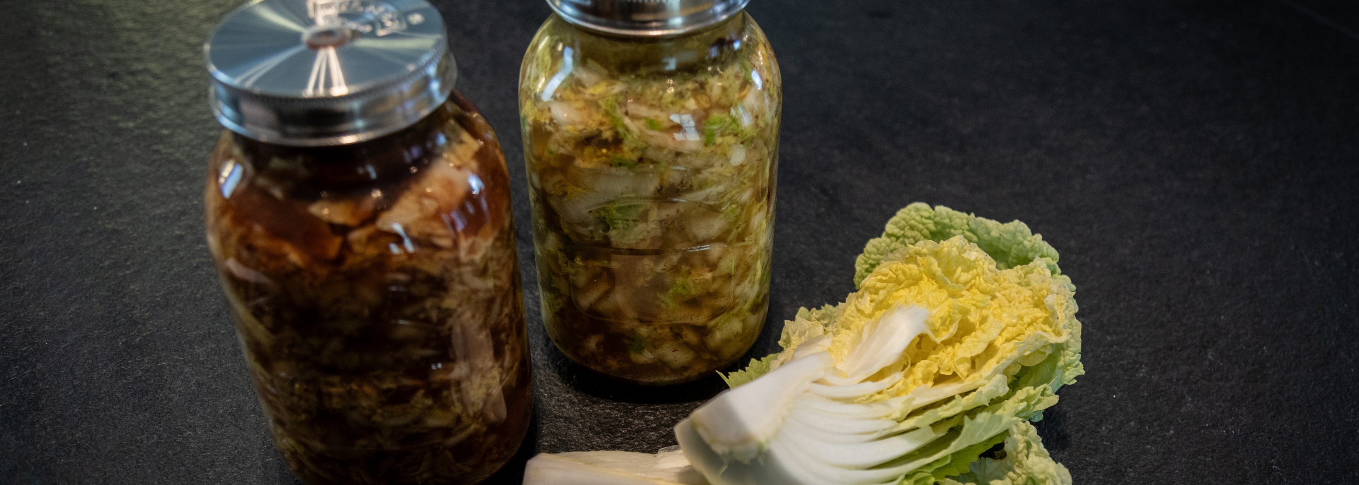 Two glass jars filled with homemade kimchi are displayed on a dark countertop beside a halved head of fresh napa cabbage, evoking precision and freshness. The setting highlights the importance of quality kitchen tools—such as Fissler’s stainless steel cookware or accessories—in preparing and storing delicious homemade creations.