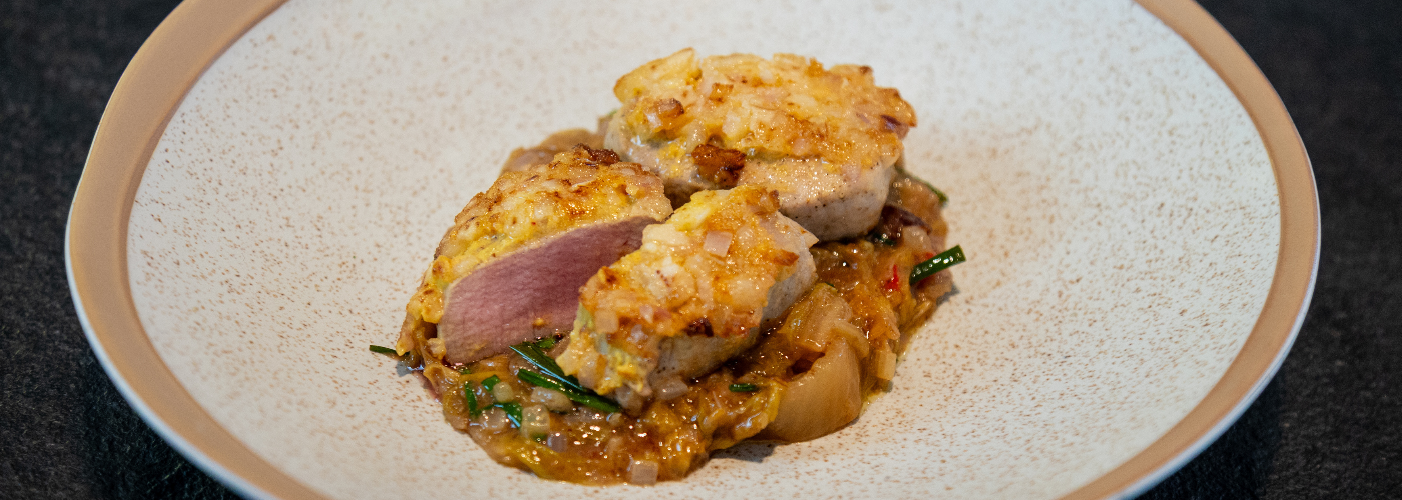 Two slices of perfectly seared pork tenderloin with a golden onion crust rest atop savory, saucy vegetables, all elegantly arranged in a speckled shallow bowl—showcasing the precision and even cooking results achieved with Fissler’s premium stainless-steel cookware.