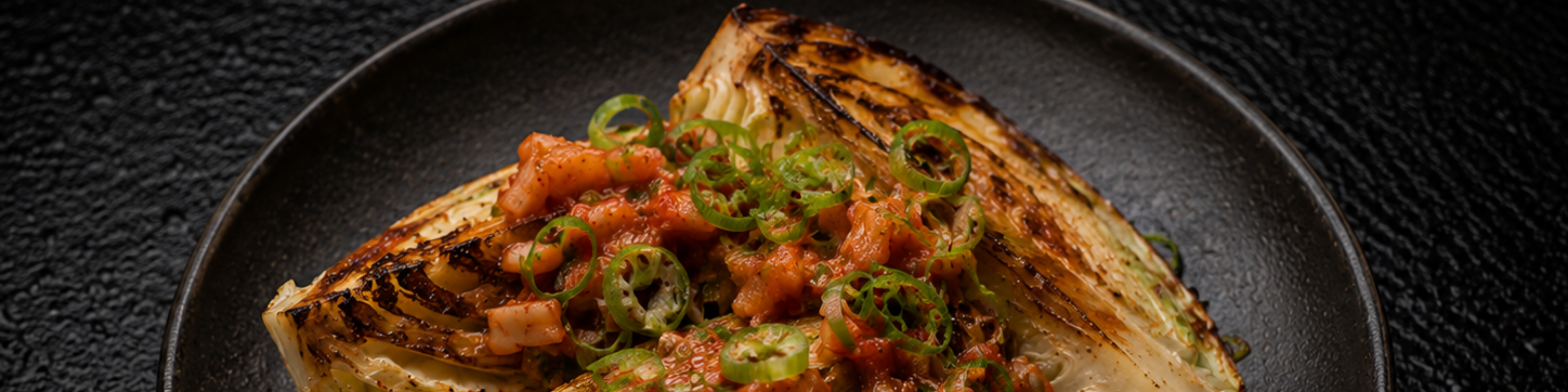 Grilled cabbage wedge topped with diced tomatoes and sliced green chili peppers, elegantly arranged on a dark plate against a textured black background. The dish, prepared using premium Fissler cookware, reflects the brand’s commitment to precision engineering and timeless German design.
