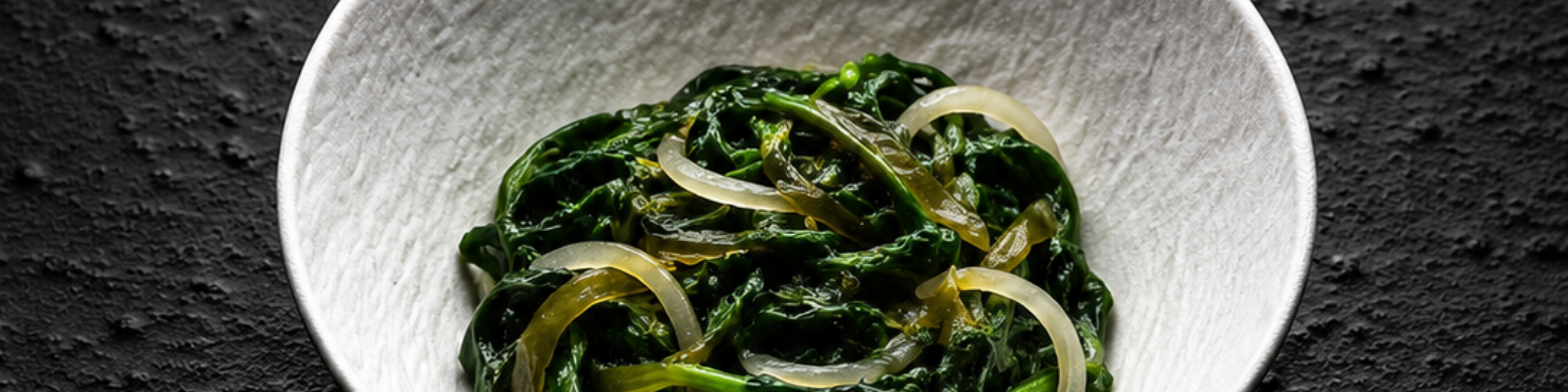 A white bowl filled with fresh seaweed salad and thinly sliced onions rests elegantly on a textured black surface, reflecting Fissler’s commitment to timeless design and quality presentation—perfect alongside our expertly crafted stainless steel cookware.