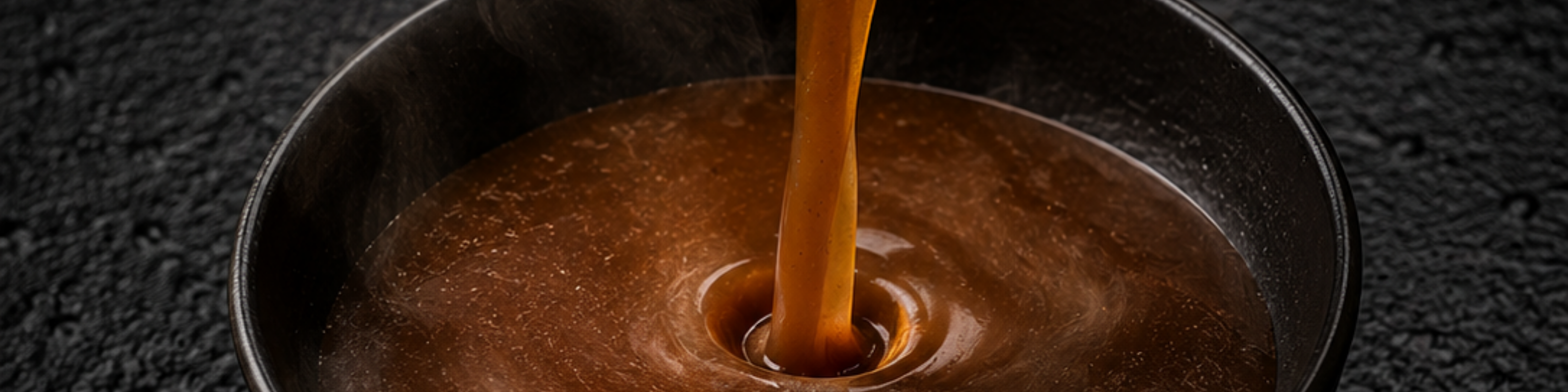 A stream of rich brown broth, simmered to perfection in a Fissler stainless steel pot, is poured into a sleek black bowl, creating elegant ripples and sending up delicate wisps of steam—a reflection of precision German engineering and timeless design.