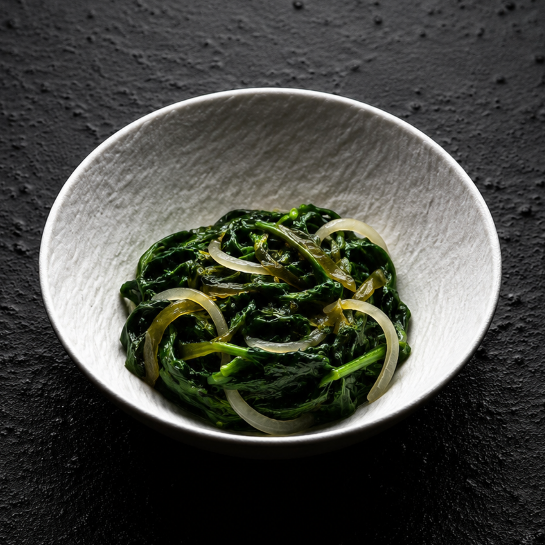 A white bowl elegantly showcases tender cooked spinach and delicately sliced onions, presented on a textured black surface—a perfect accompaniment to gourmet dishes prepared with Fissler’s precision-engineered cookware, reflecting our timeless German craftsmanship.