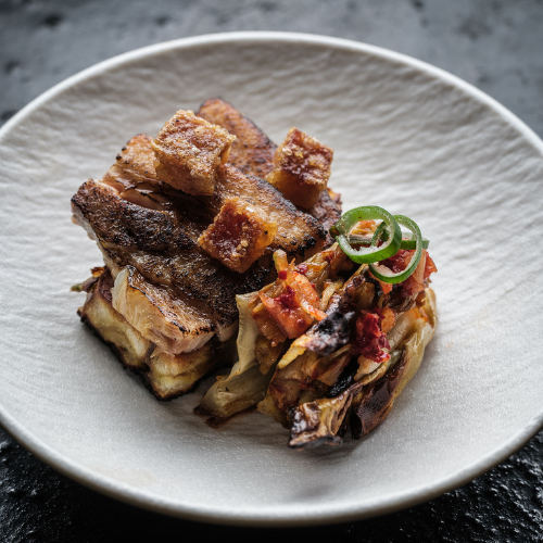 Crispy pork belly with golden crackling, elegantly presented alongside roasted vegetables and garnished with fresh green herbs, served on a refined white plate—expertly prepared using Fissler’s precision-engineered cookware for outstanding culinary results.