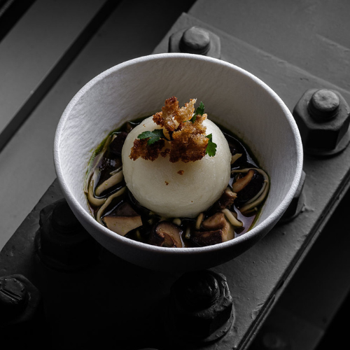 A meticulously plated dish in a white bowl, featuring a round, white scoop garnished with crispy topping, nestled atop sliced mushrooms and fresh herbs in a rich dark broth. The presentation sits on an industrial metal surface with visible bolts—a nod to Fissler’s precision-engineered stainless steel cookware "Made in Germany," blending culinary artistry with timeless, durable German design.