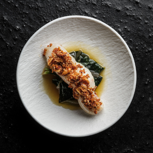 A flawlessly presented piece of fish with a crispy garnish is placed atop fresh greens and complemented by a delicate light brown sauce, all arranged on a pristine white plate. The elegant dish sits on a textured black surface—reflecting the precision and timeless design associated with Fissler’s expertly crafted cookware.