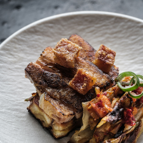 A close-up of a beautifully arranged dish featuring crispy pork belly cubes stacked atop expertly grilled meat, accompanied by roasted vegetables and sliced green onions, elegantly presented on a white plate—prepared and served with precision using Fissler’s premium stainless steel cookware.