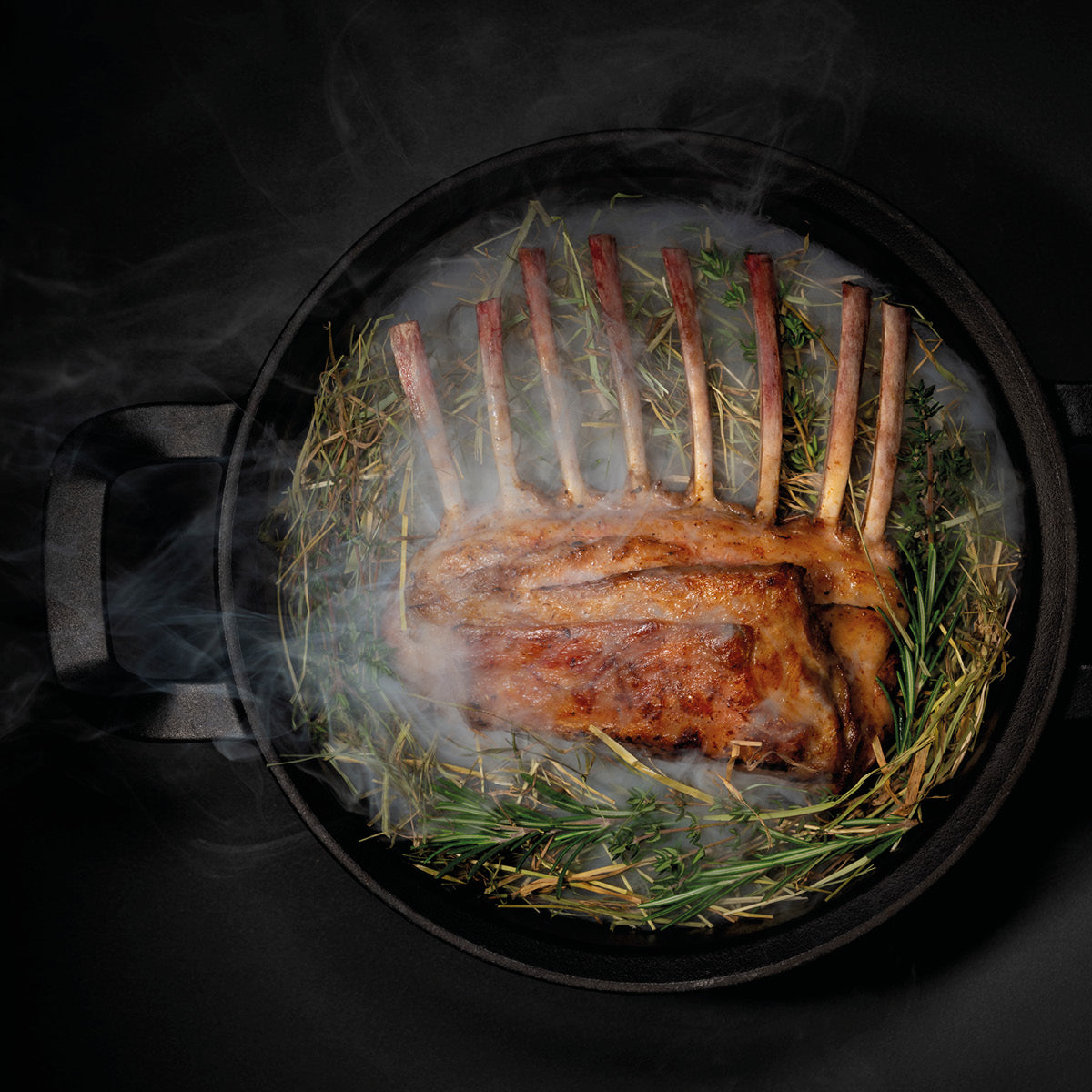 Ein Lammrücken auf Kräutern und Heu in einer Fissler Gusseisenpfanne gebraten, sanfter Rauch steigt auf - Präzision und Qualität "Made in Germany".