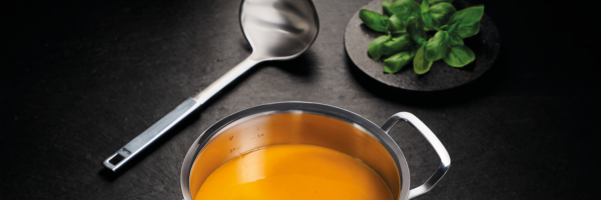 Ein Fissler-Edelstahltopf mit orangefarbener Cremesuppe, daneben eine silberne Schöpfkelle und frisches Basilikum - das ist deutsches Qualitätsdesign.