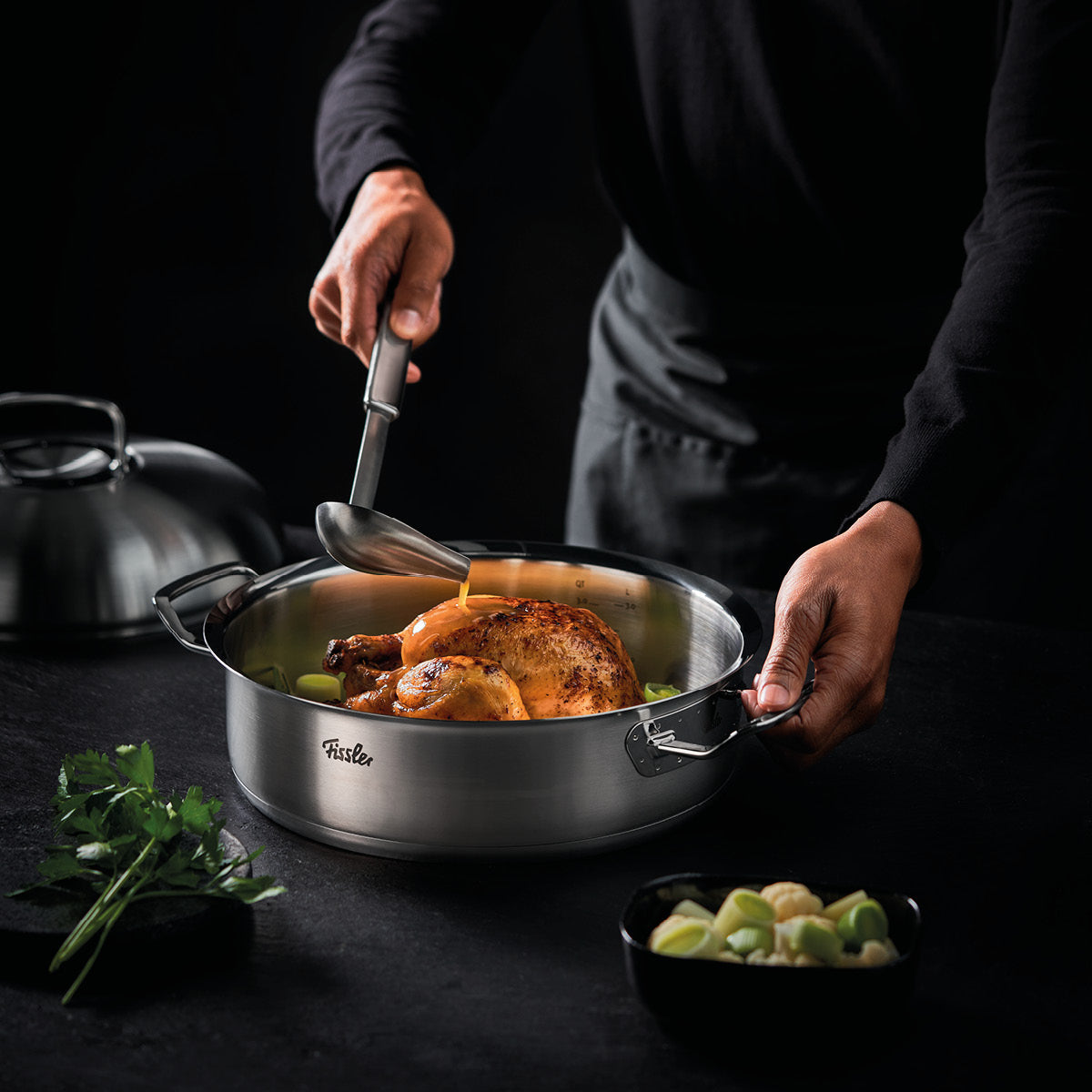 Eine Person brät ein Hähnchen in einem Fissler-Edelstahltopf; frische Kräuter und Gemüse liegen auf einem schlichten dunklen Tisch daneben.