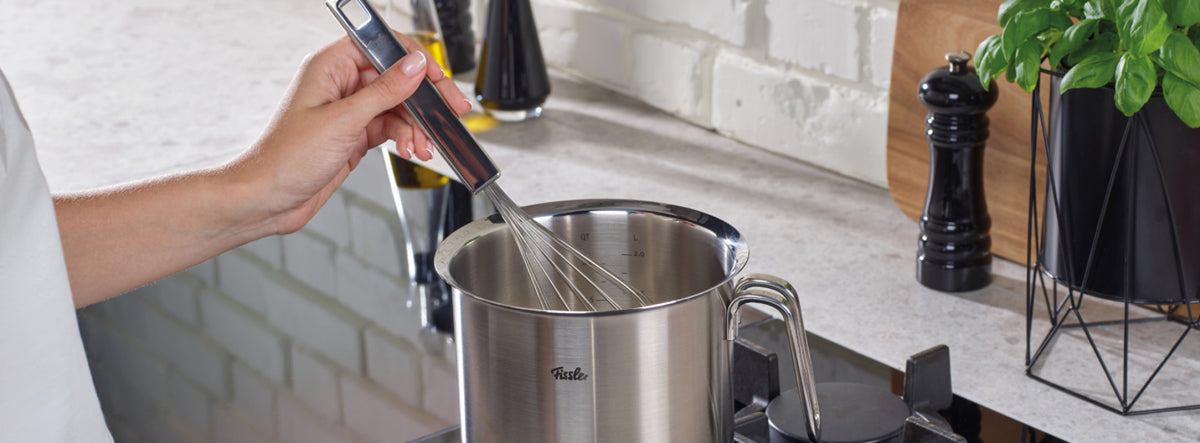 Eine Person rührt Zutaten in einem hochwertigen Fissler-Edelstahltopf auf dem Herd um, während in der Nähe hochwertige Küchengeräte elegant angeordnet sind.
