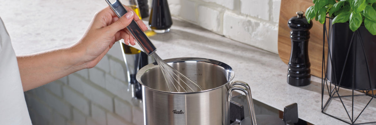 Eine Person kocht in einem Fissler-Edelstahltopf auf dem Herd, während im Hintergrund hochwertige Küchenutensilien und frisches Basilikum zu sehen sind.