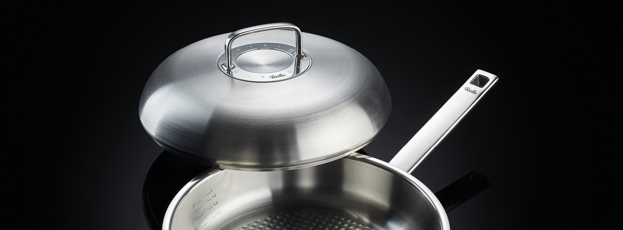 Ein Fissler-Edelstahltopf mit langem Griff und gewölbtem Deckel, teilweise geöffnet, präsentiert auf einer spiegelnden schwarzen Oberfläche.