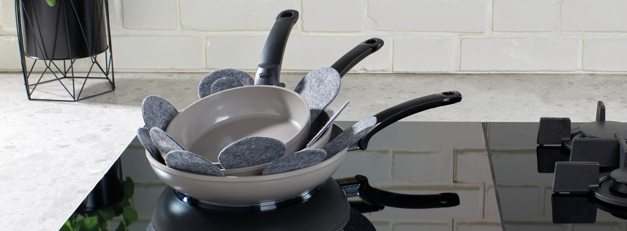 Fissler Antihaft-Bratpfannen mit ergonomischen schwarzen Griffen, gestapelt auf einem Herd mit Filzschutz; helle Backsteinplatte sichtbar.