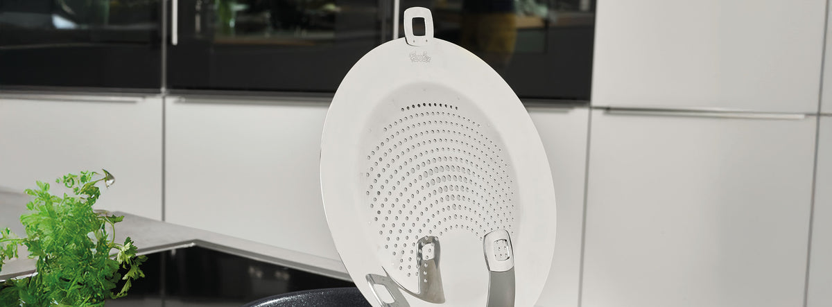 Ein weißer Silikon-Spritzschutz von Fissler ruht auf einer schwarzen Pfanne in einer modernen Küche mit Geräten aus Edelstahl und frischen Kräutern.