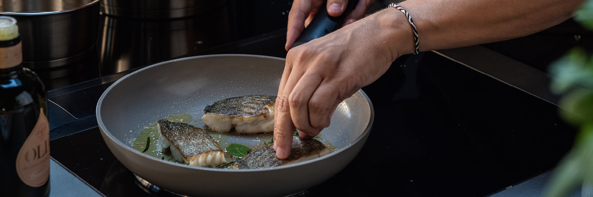 Die Hände braten fachmännisch zwei mit Kräutern gewürzte Fischfilets in einer Fissler-Pfanne, wobei das hochwertige Kochgeschirr auf der Herdplatte sichtbar bleibt.