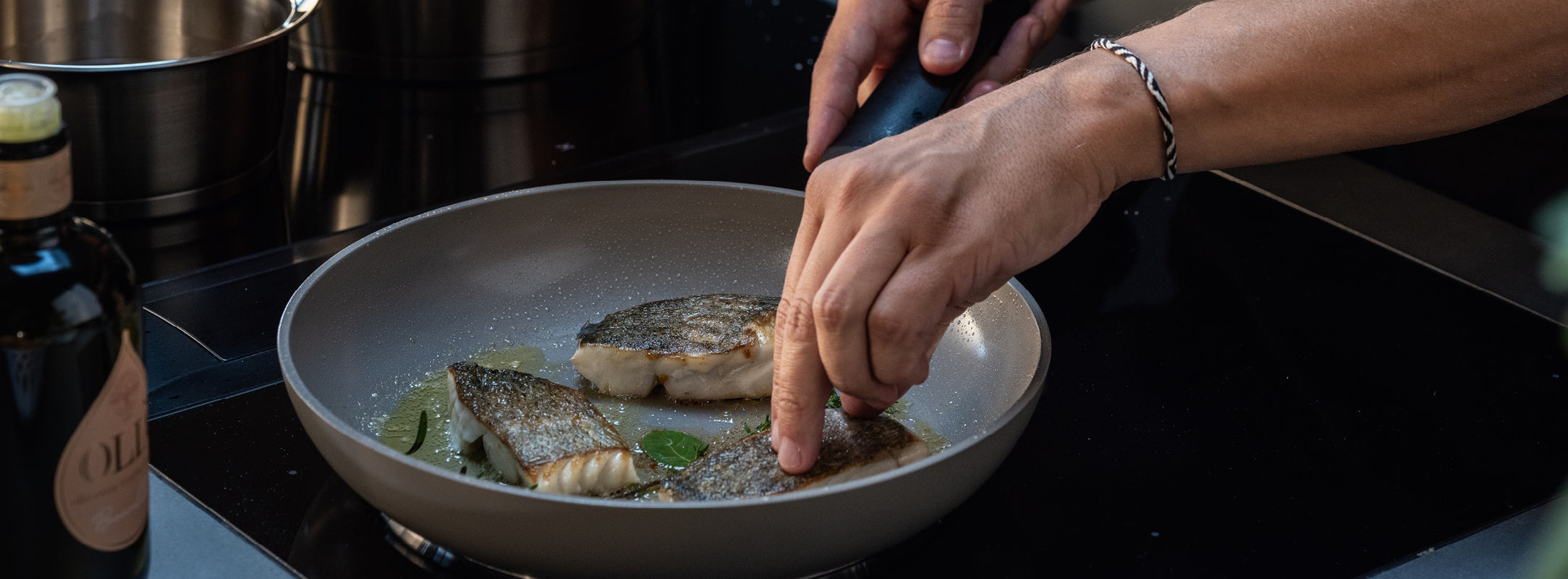 Ein Koch benutzt eine Fissler-Zange und eine hochwertige Bratpfanne, um Fischfilets mit Kräutern auf dem Herd zu braten. Eine Flasche Olivenöl steht daneben.