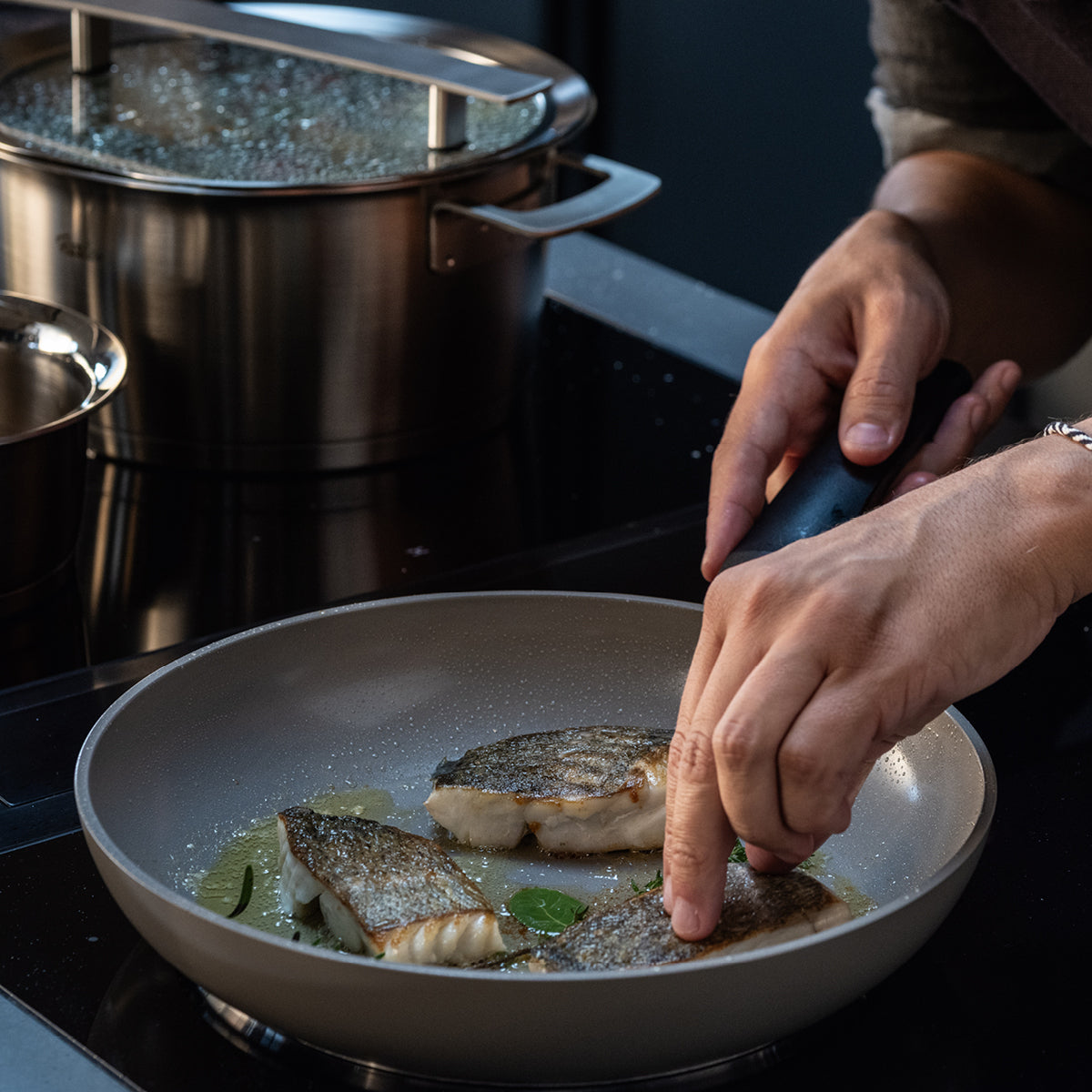 Ein Koch richtet Fischfilets in einer Fissler-Pfanne mit Öl und Kräutern an; daneben steht ein abgedeckter Fissler-Topf auf der Herdplatte.