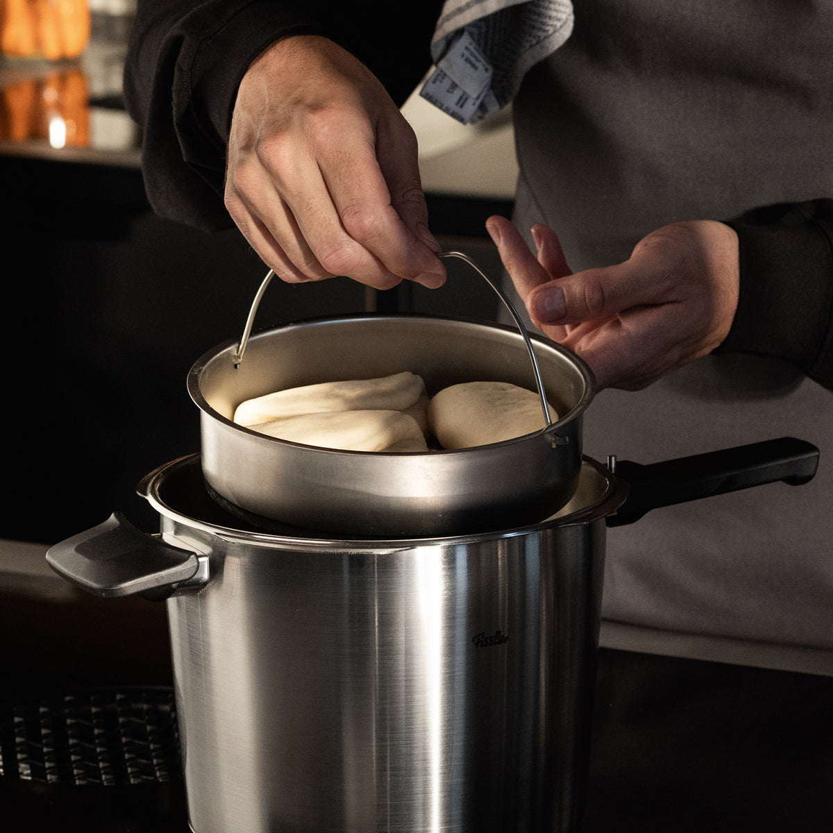 Eine Person hebt einen Dämpfkorb mit drei rohen Brötchen aus einem Fissler-Edelstahltopf, der von deutscher Präzision und Qualität zeugt.