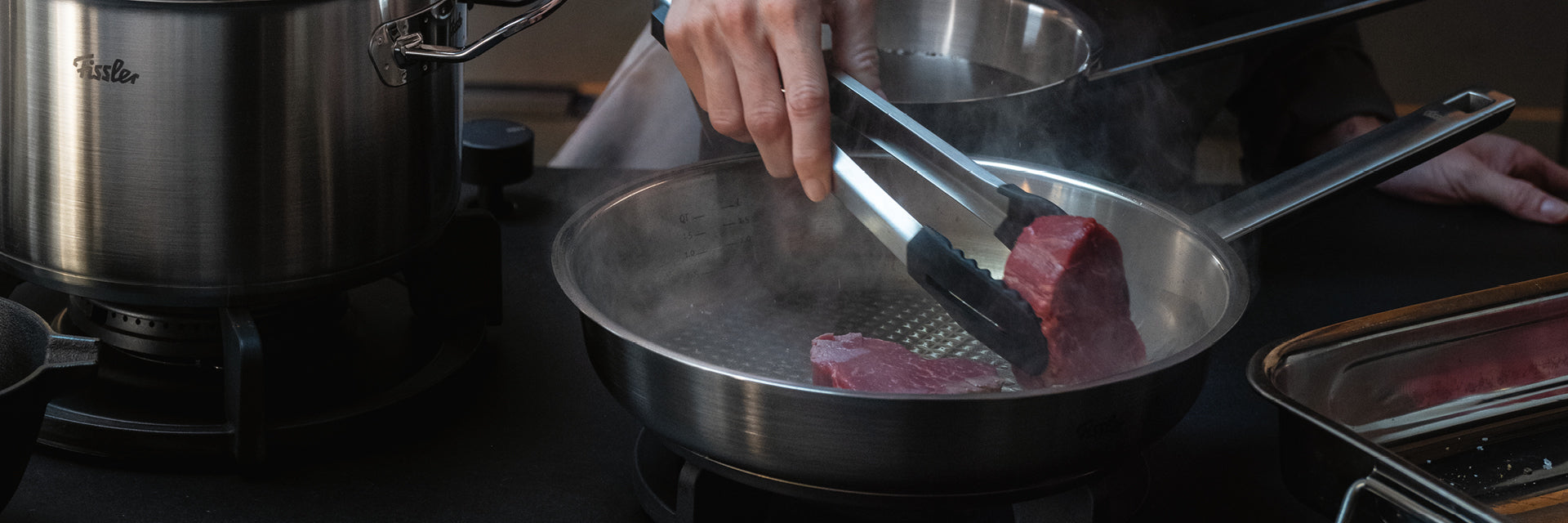 Eine Person brät ein Steak mit einer Zange in einer hochwertigen Fissler-Edelstahlpfanne an; elegante Töpfe und Pfannen glänzen auf der Küchentheke.