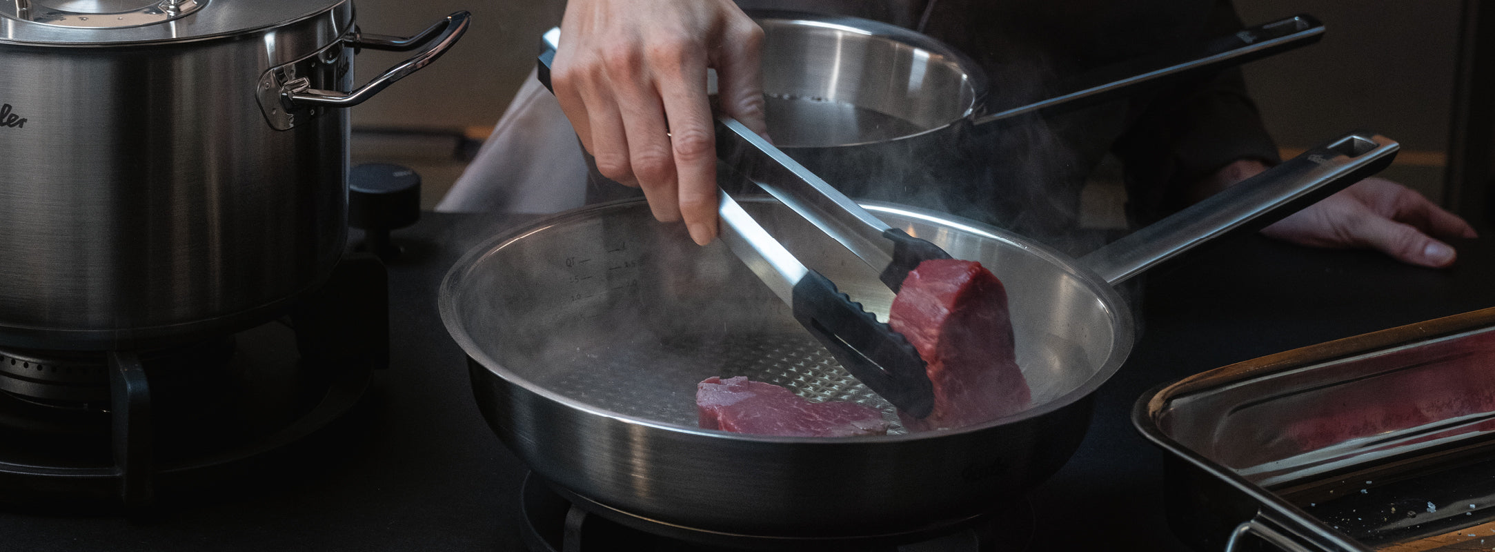 Eine Person benutzt eine Zange, um ein rohes Steak in eine hochwertige Fissler-Edelstahlpfanne auf einem Herd zu legen, Kochgeschirr in der Nähe.