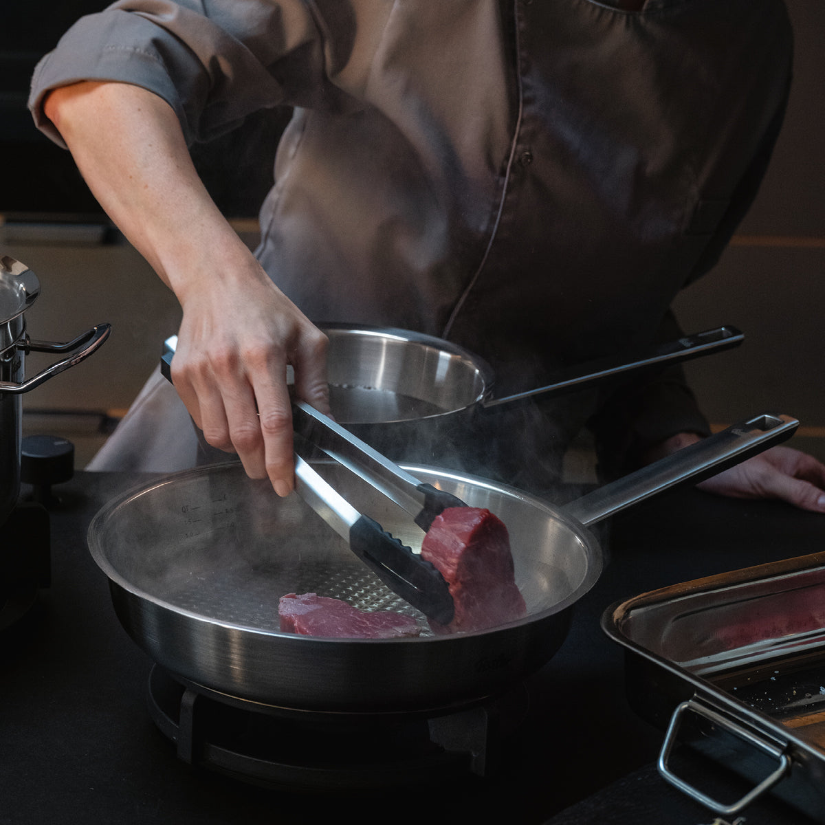 Ein Koch legt ein rohes Steak in eine Fissler-Edelstahlpfanne auf dem Herd, neben der weiteres hochwertiges Fissler-Kochgeschirr steht.