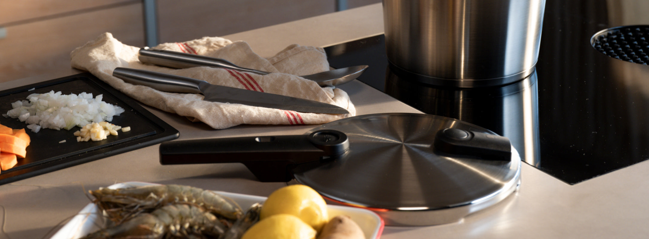 Ein Fissler-Edelstahltopf auf dem Herd, ein Schnellkochtopfdeckel, Messer und frische Zutaten auf einer modernen Küchentheke.