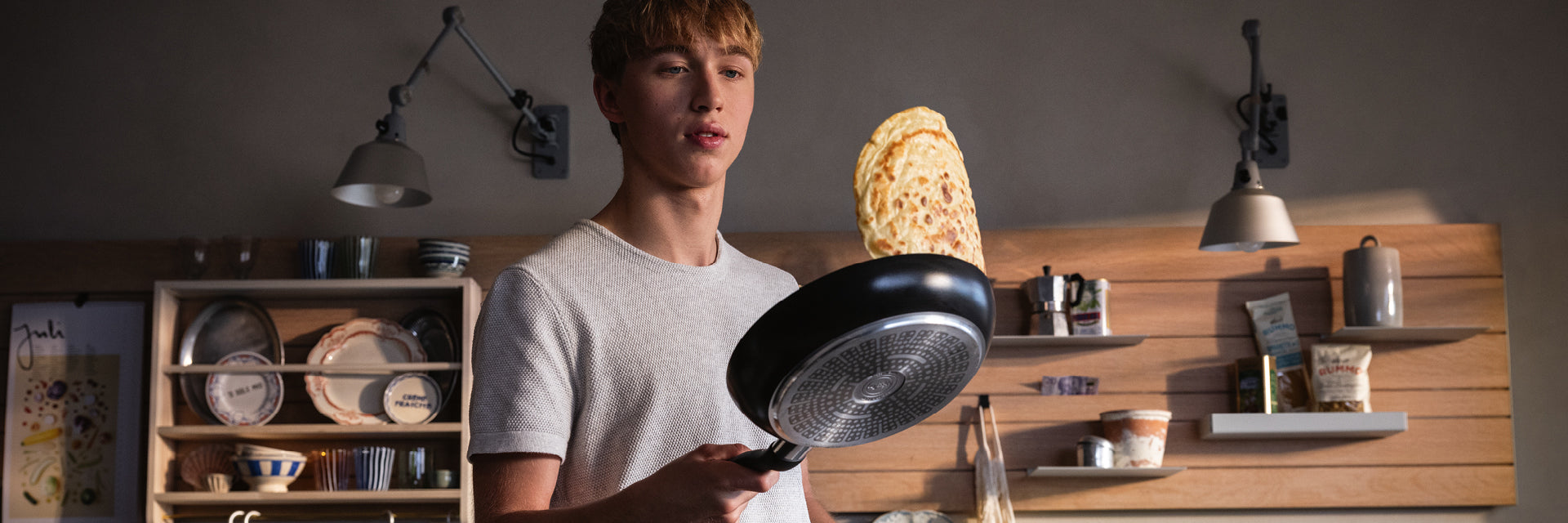 Eine junge Person in einem grauen T-Shirt wendet einen Pfannkuchen in einer Fissler-Pfanne in einer modernen Küche mit sichtbaren Regalen und Kochgeschirr.