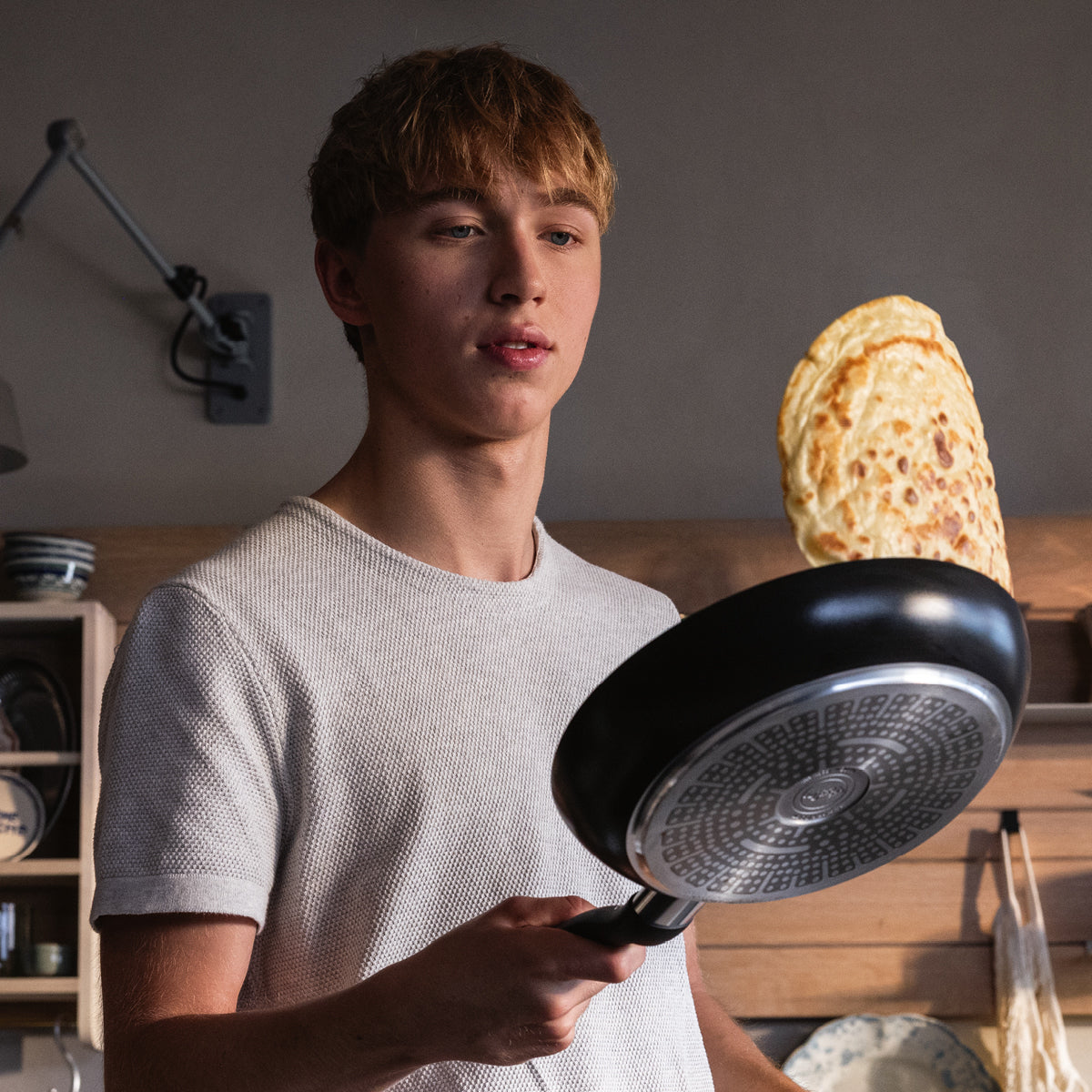 Junger Mann wendet einen Pfannkuchen in einer Fissler-Pfanne, konzentriert in einer modernen Küche mit Regalen voller Teller und Schüsseln im Hintergrund.