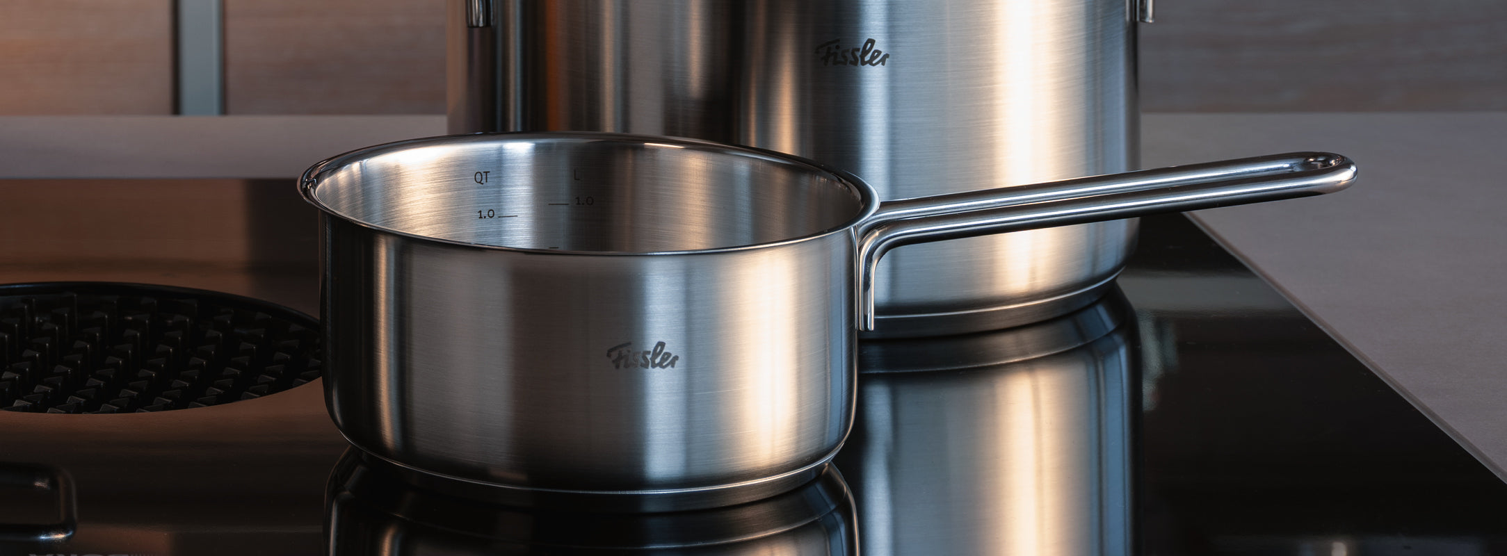 Fissler-Topf aus rostfreiem Stahl mit langem Griff auf einer schwarzen Glas-Kochplatte. Im Hintergrund ein weiterer Fissler-Topf, beide mit Logo.