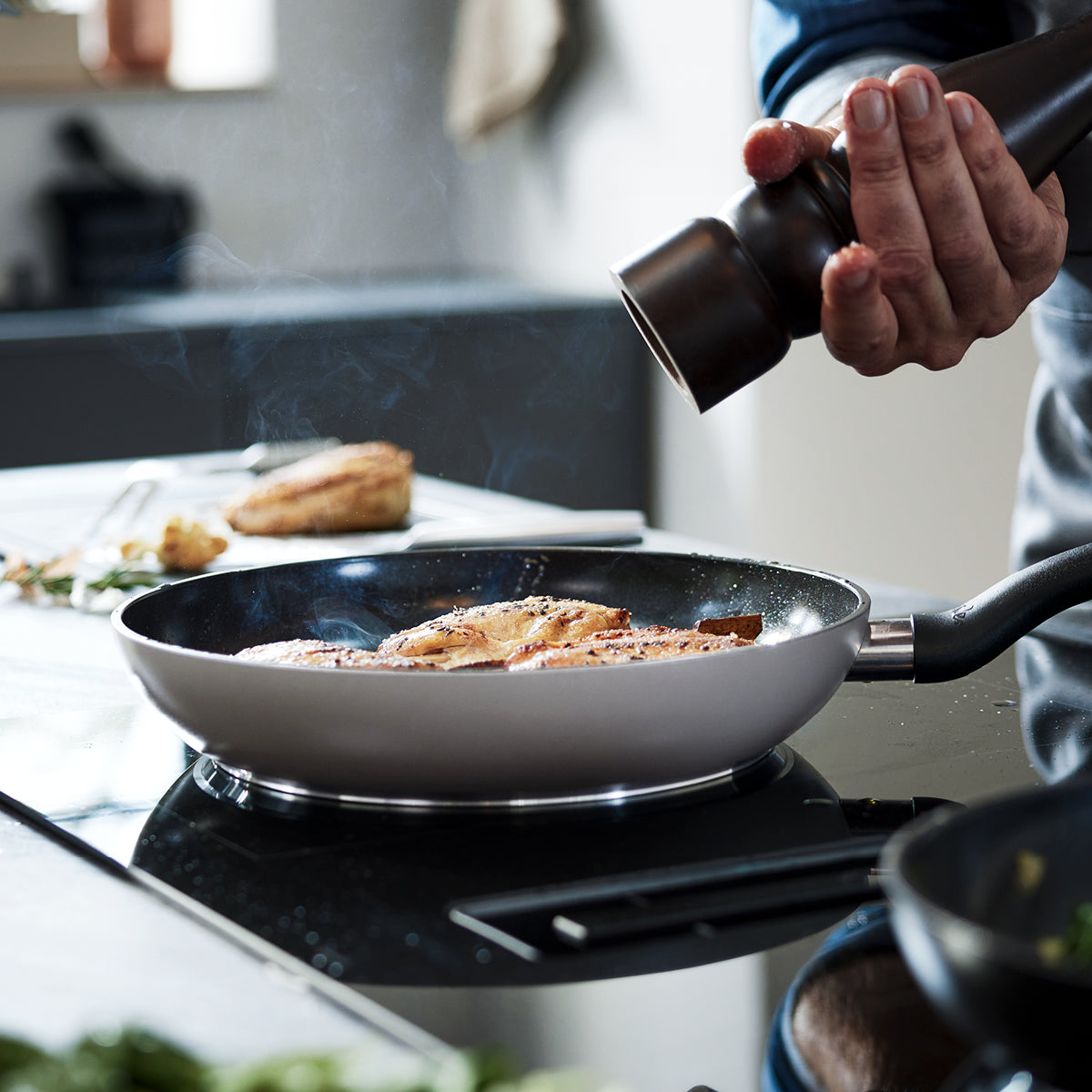 Eine Person würzt Fischfilets, die in einer hochwertigen Fissler-Edelstahlpfanne garen, während in einer modernen Küche Dampf aufsteigt.