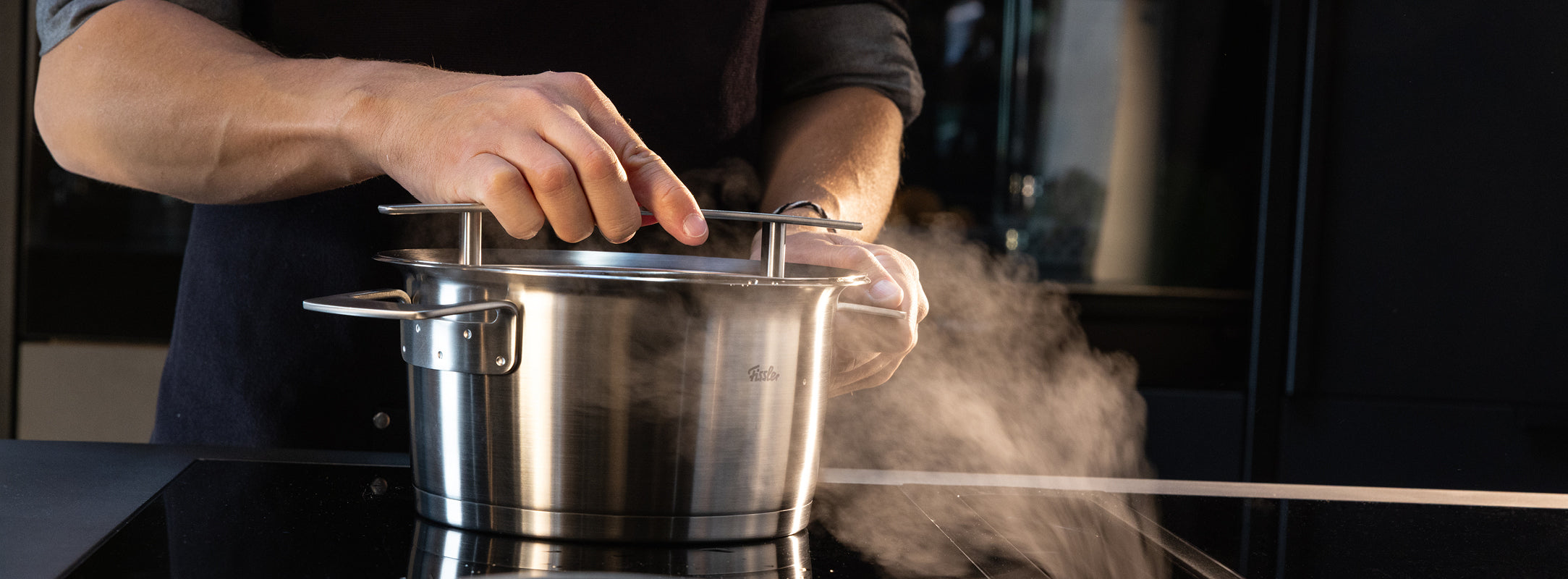 Eine Person hebt den Deckel eines Fissler-Edelstahltopfes an und präsentiert deutsche Technik und aufsteigenden Dampf auf einer modernen Herdplatte.