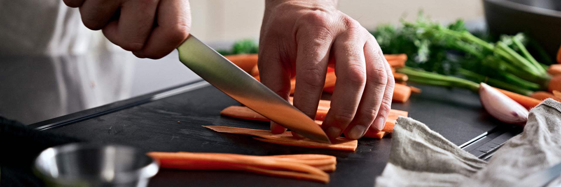 Nahaufnahme von Händen, die mit Präzision Karotten auf einem Schneidebrett schneiden, umgeben von frischem Obst und Gemüse - wie geschaffen für die Fissler-Küche.