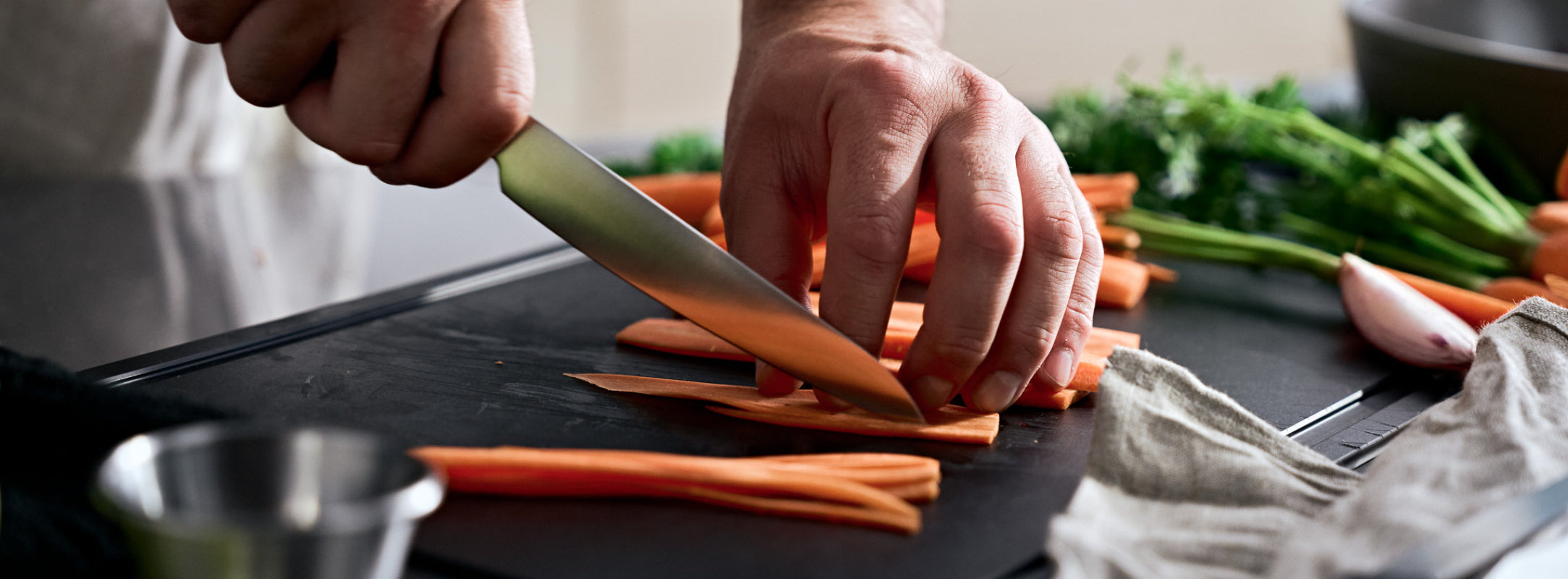 Eine Person schneidet Karotten auf einem Fissler-Schneidebrett, umgeben von hochwertigem Kochgeschirr und frischem Gemüse in einer modernen Küche.