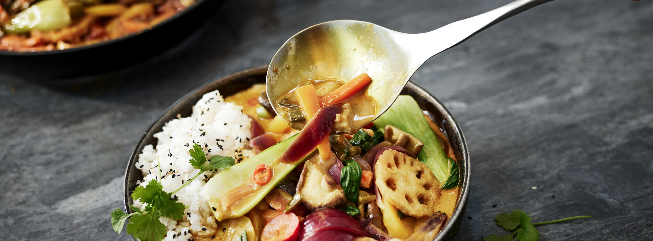 Ein lebhaftes Gemüsecurry mit Reis, serviert in einer Fissler-Schüssel. Ein Löffel hebt das Curry auf und zeigt die Exzellenz des deutschen Kochgeschirrs.