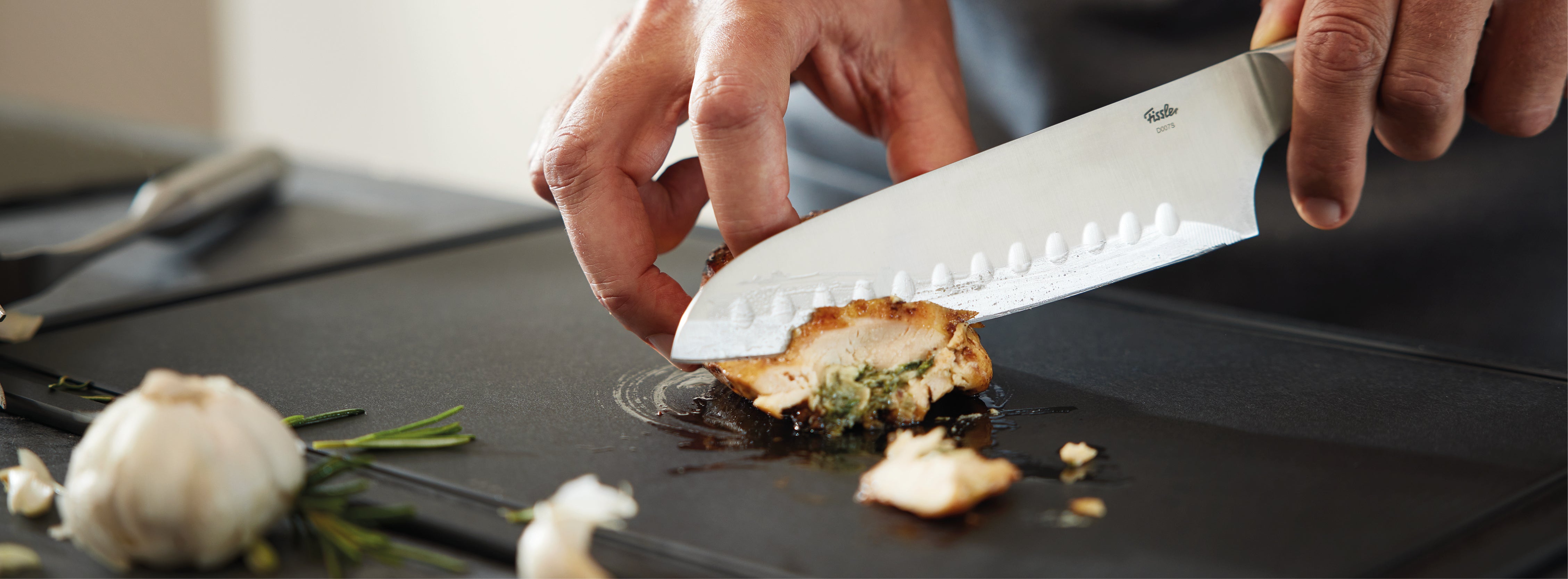 Eine Hand schneidet die gefüllte Hühnerbrust auf einem Fissler-Schneidebrett präzise in Scheiben, wobei Knoblauch und Rosmarin für kulinarische Inspiration sorgen.