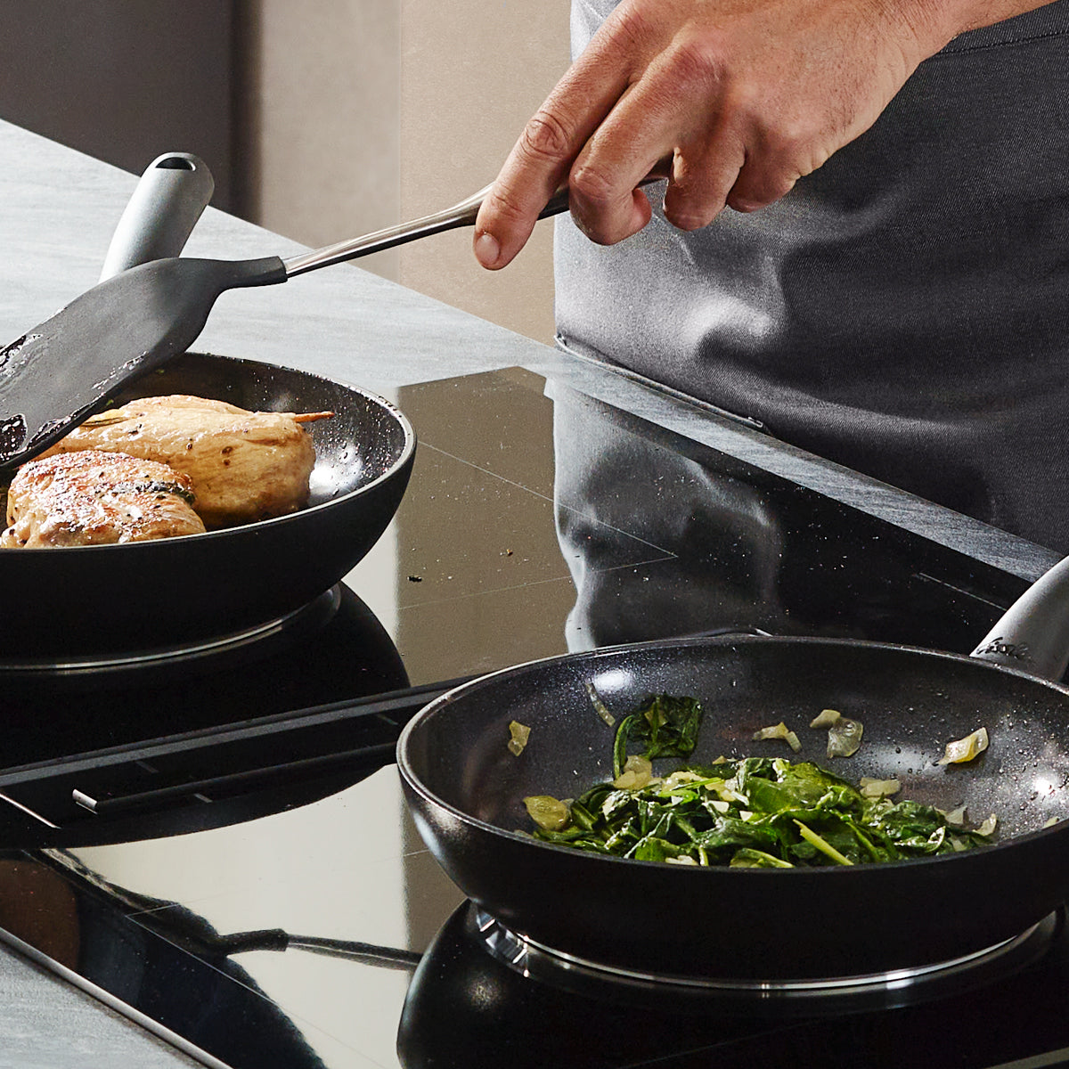 Eine Person kocht Hähnchen in einer Fissler-Pfanne und Grünzeug in einer anderen, mit einem Fissler-Pfannenwender auf einem eleganten, in Deutschland entwickelten Herd.