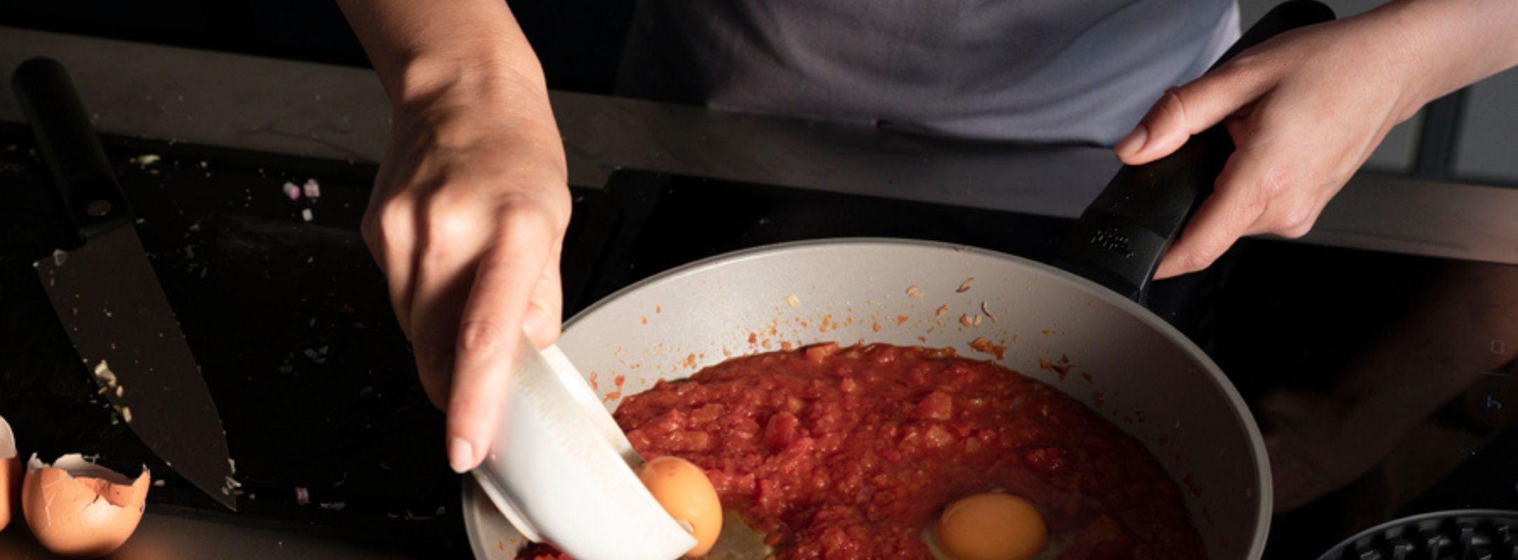 Eine Person schlägt ein Ei in eine Fissler-Pfanne mit Tomatensauce auf dem Herd auf; Eierschalen und ein Messer liegen auf dem Tresen daneben.
