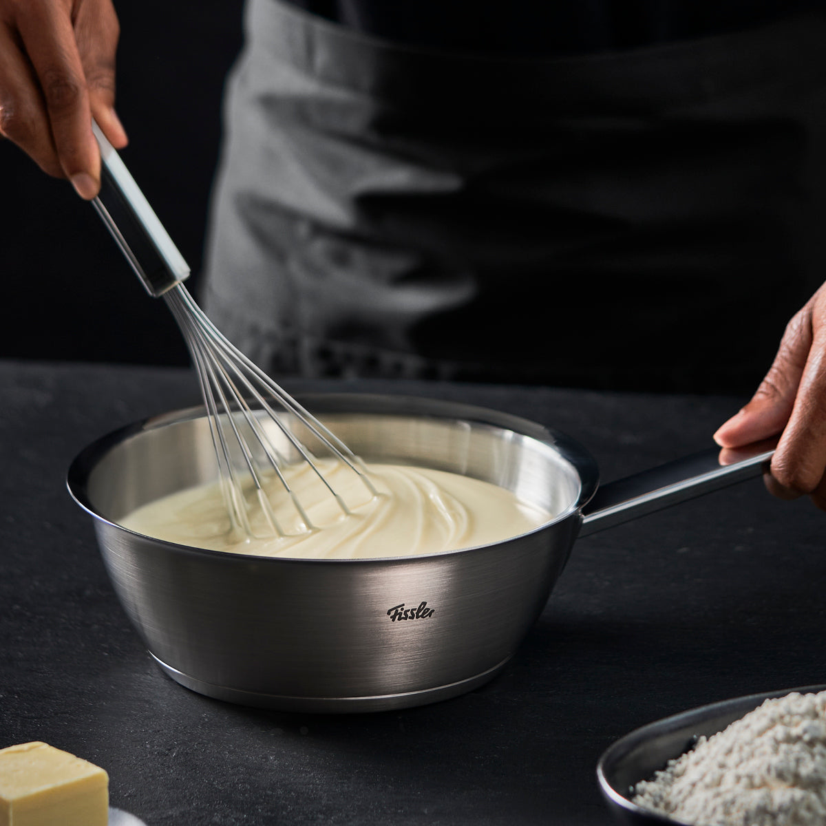Eine Person rührt eine cremige Masse in einem Fissler-Edelstahltopf, der deutsche Präzision und zeitloses Design demonstriert. Mehl und Butter in der Nähe.