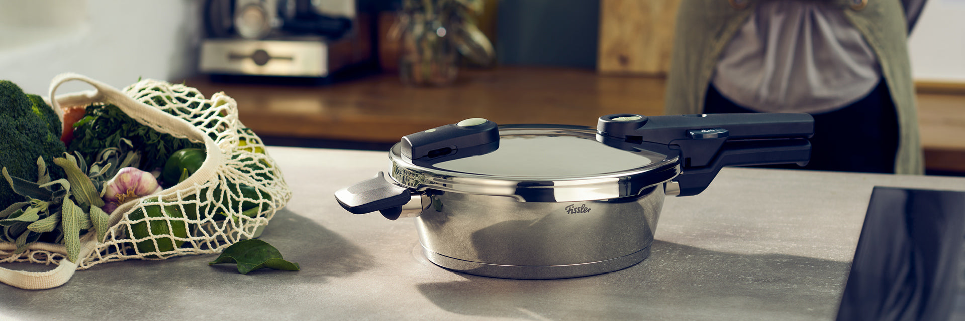 Ein Fissler Schnellkochtopf aus rostfreiem Stahl, Deckel geschlossen, auf einem Tresen neben einem Netzbeutel mit Gemüse; Person unscharf im Hintergrund.