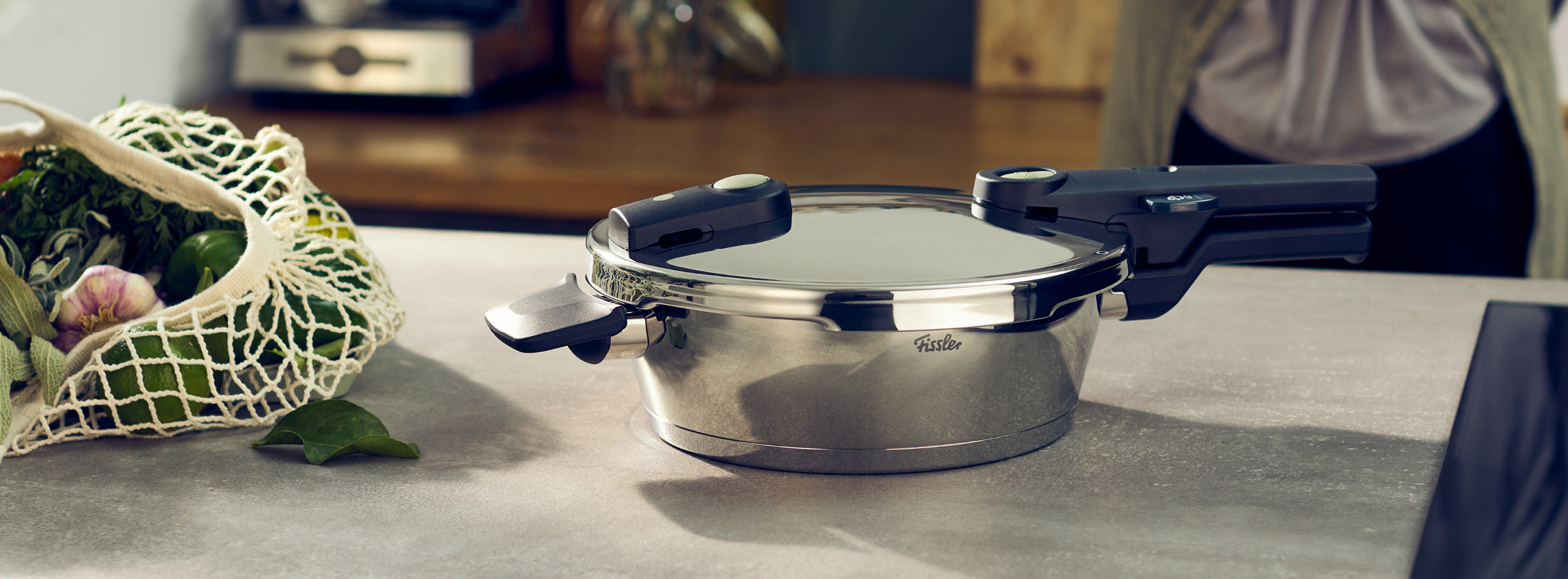 Ein Fissler Schnellkochtopf aus rostfreiem Stahl mit schwarzen Griffen steht auf einer Küchenzeile neben frischem Gemüse. Dahinter steht eine Person.