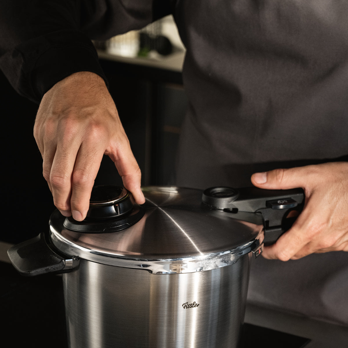 Eine Person in dunklem Hemd und Schürze verstellt den Deckel und den Drehknopf eines Fissler Schnellkochtopfes aus Edelstahl, Made in Germany.