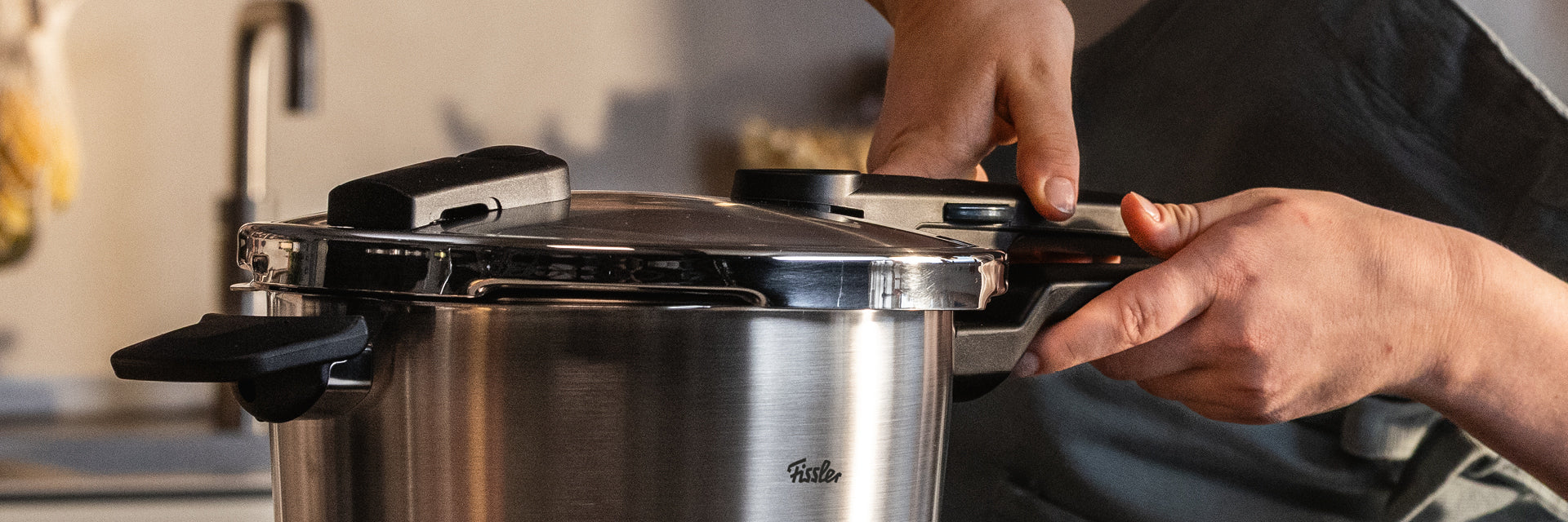 Die Hände schließen sicher den Deckel eines Fissler Schnellkochtopfes aus Edelstahl, der mit deutscher Präzision und zeitlosem Design in der Küche glänzt.