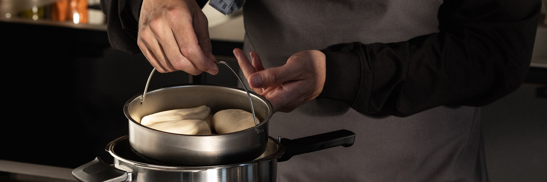 Eine Person in dunklem Hemd und Schürze hebt einen Fissler-Edelstahltopf mit gedünsteten Brötchen über einen passenden Kochtopf auf dem Herd.