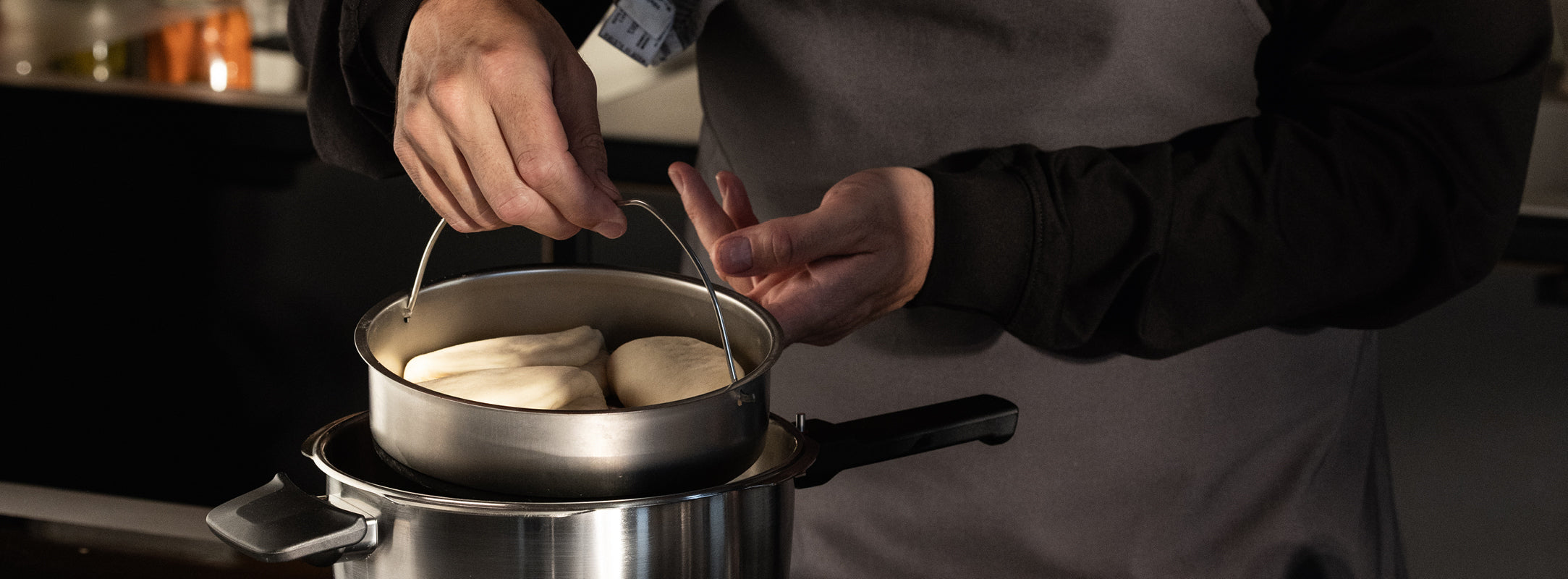 Eine Köchin in grauer Schürze verwendet gestapelte Fissler-Edelstahltöpfe zum Dämpfen von Teig, die Präzision und zeitloses deutsches Design demonstrieren.